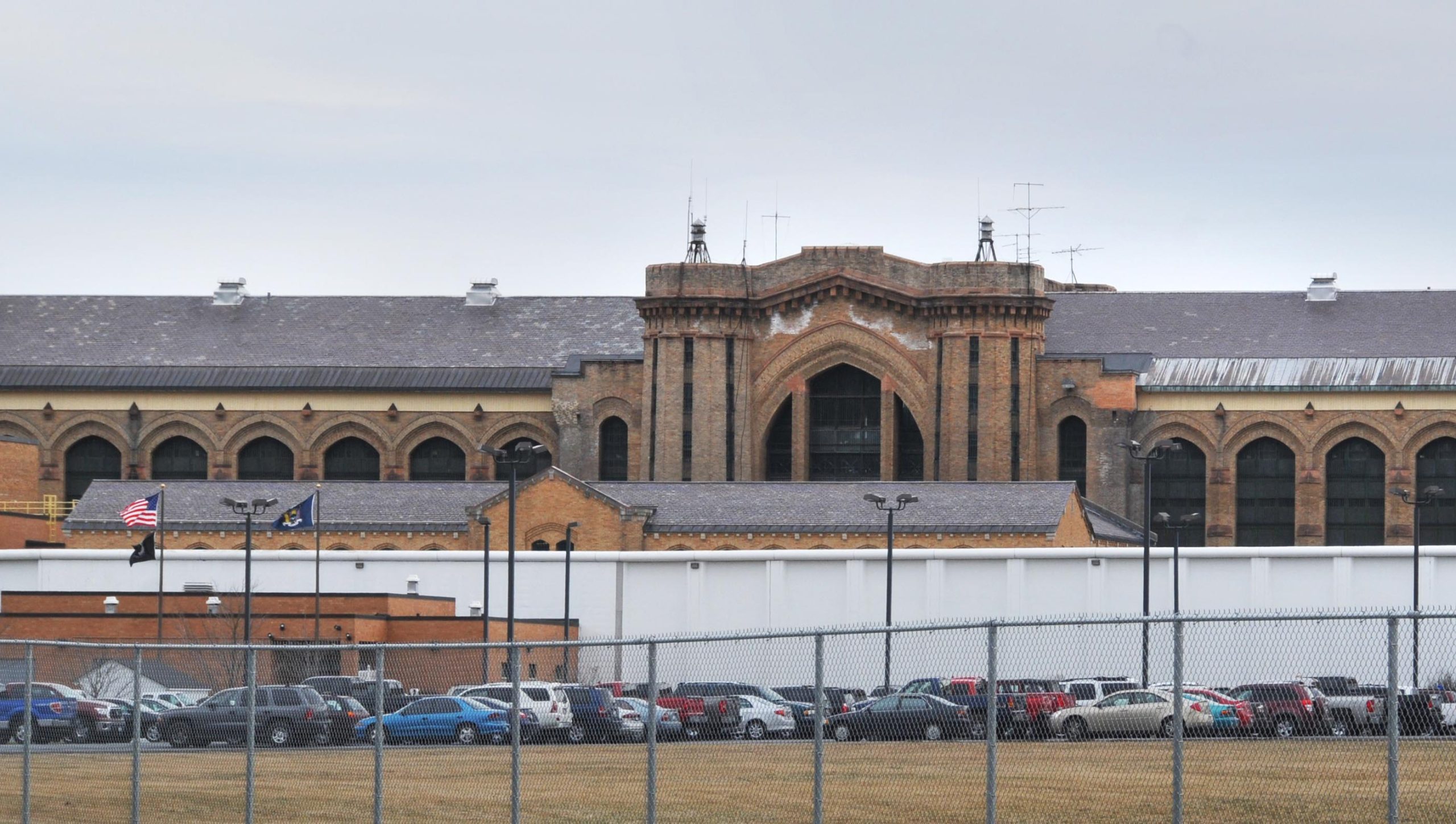 A view of the exterior of Great Meadow Correctional Facility during the day. Cars are parked in front of the orange-brown colored building, which has arched windows. 